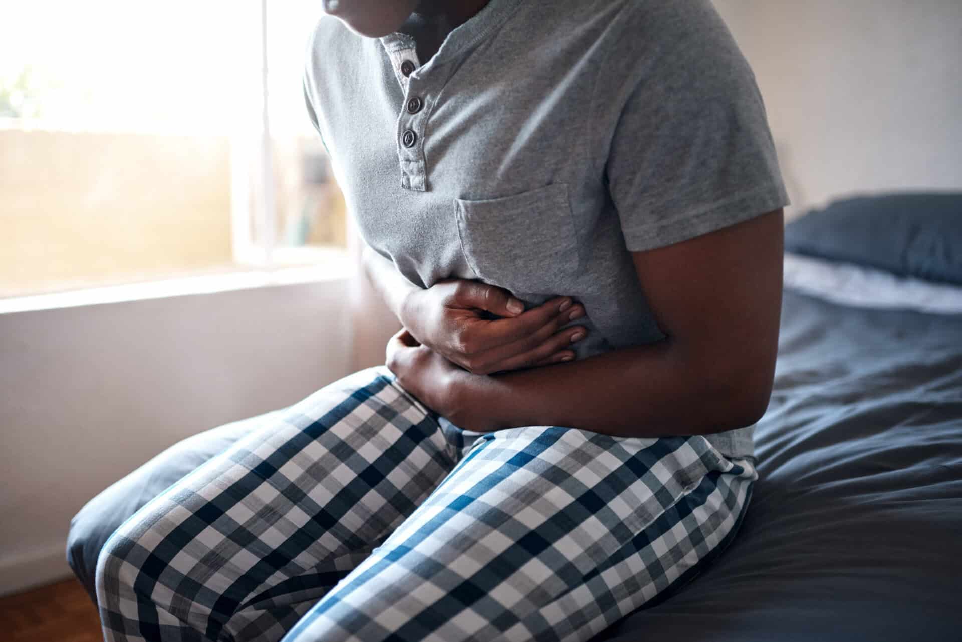 Man, stomach ache and hands holding belly in a bedroom in a home at morning. African male person, sick and indigestion pain in a house on a bed feeling hurt with abdomen cramp and ibs problem.